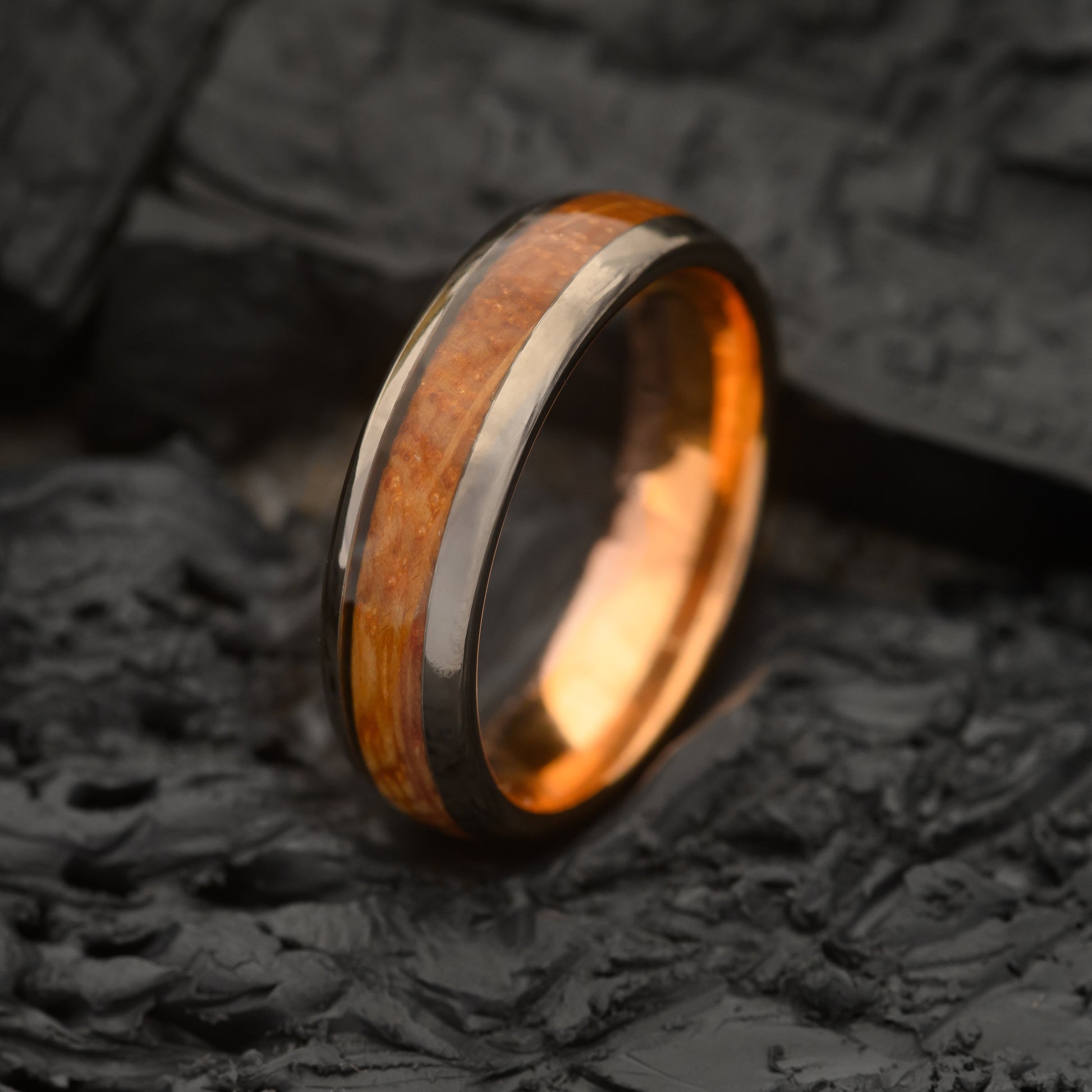 A Mens 6mm Tungsten steel ring with single groove whiskey barrel inlay and rosegold inner ring resting on Charcoal slate bed