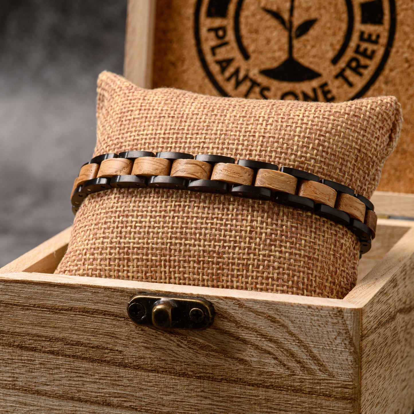 A whiskey wood and tungsten steel bracelet wrapped around a hessian cushion sitting in a wooden box with a grey stone backdrop