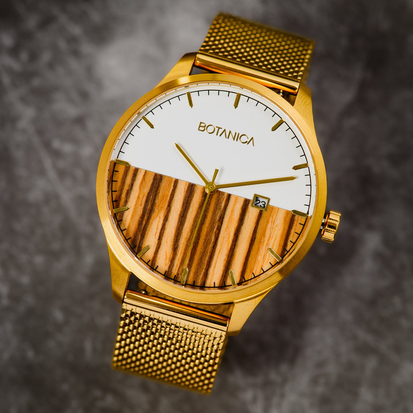 A gold stainless steel watch with a white and wood patterned dial, featuring a gold mesh band and the brand name 'BOTANICA' on the dial.