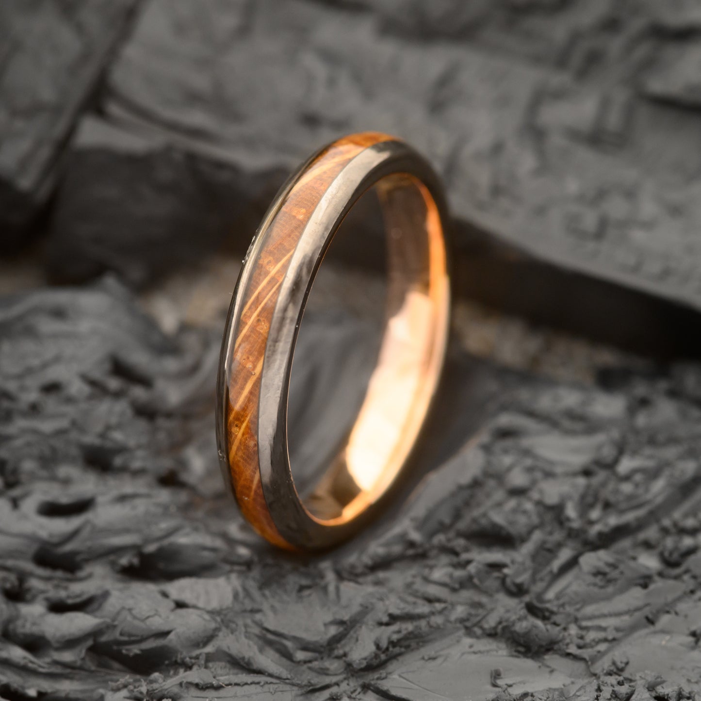 A Womens 4mm tungsten steel ring with rosegold & whiskey barrel inlay resting on a charcoal slate