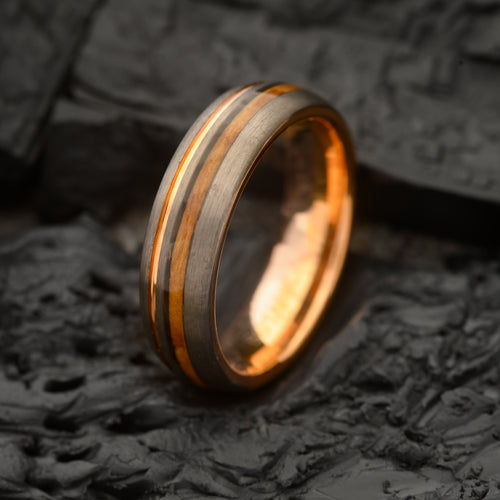 A charcoal grey tungsten steel ring with a rose gold band and a strip of wood inlay, displayed on a textured dark background.