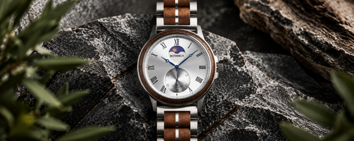A wristwatch with a silver-toned metal case, white dial, blue hands and moonphase indicator, and brown wood-and-metal link band, photographed resting on dark rocks with blurred greenery around it.