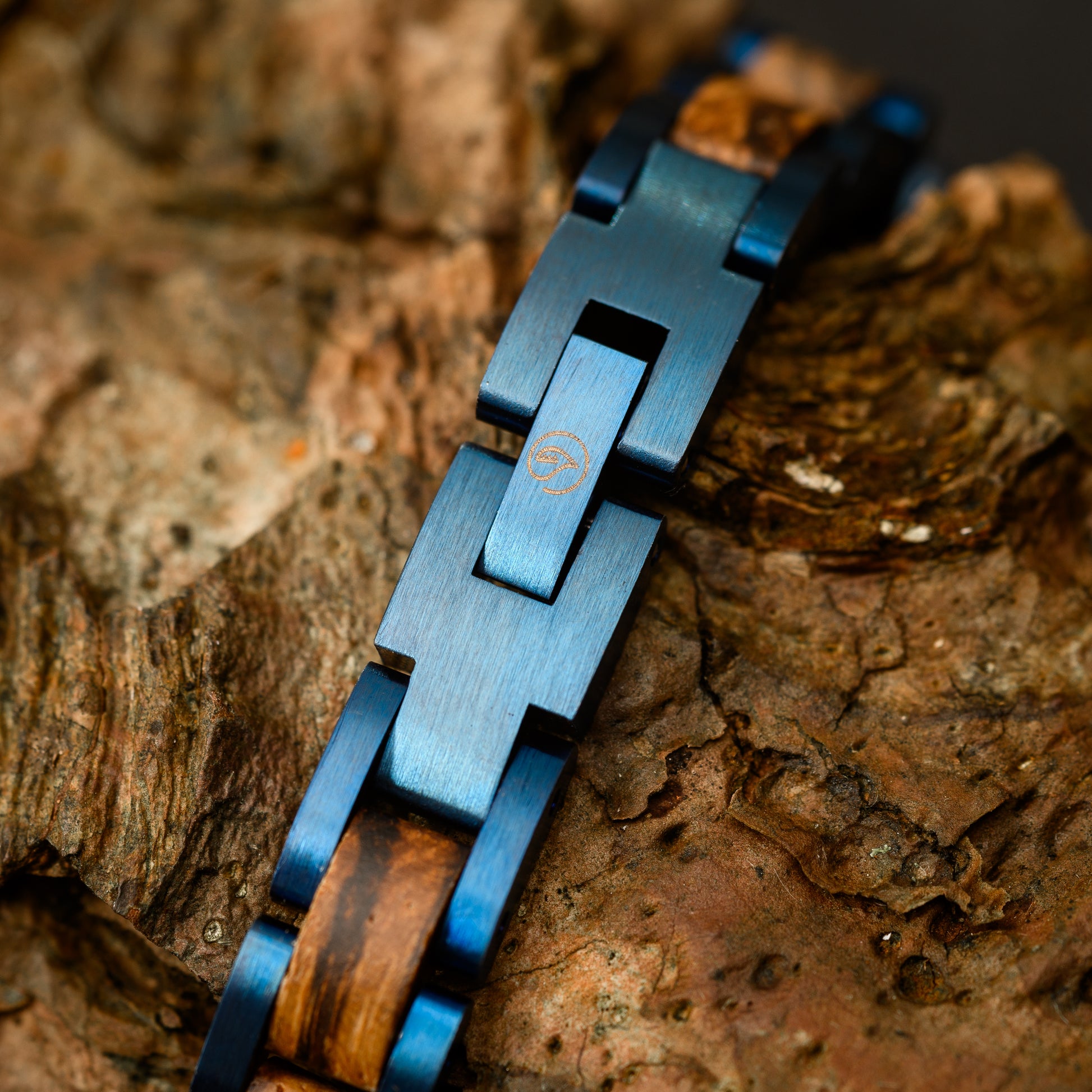 A close up detail of the Botanica Logo clasp on the blue tungsten steel and zebrano wood bracelet resting on drift wood