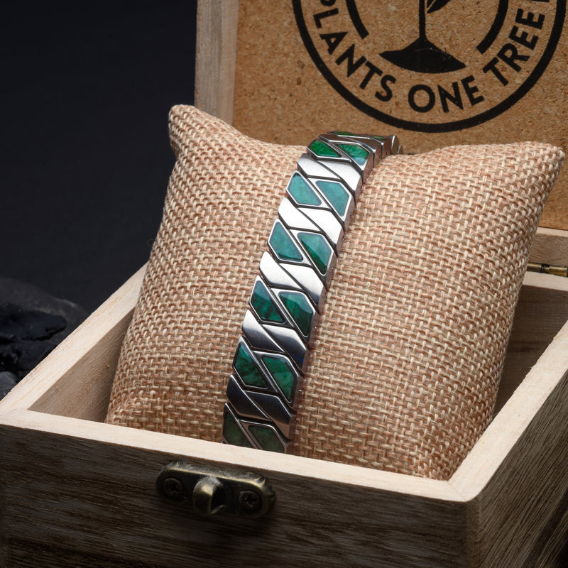 Bracelet with green and silver pattern on a woven cushion in a wooden box, branded 'Plants One Tree'.