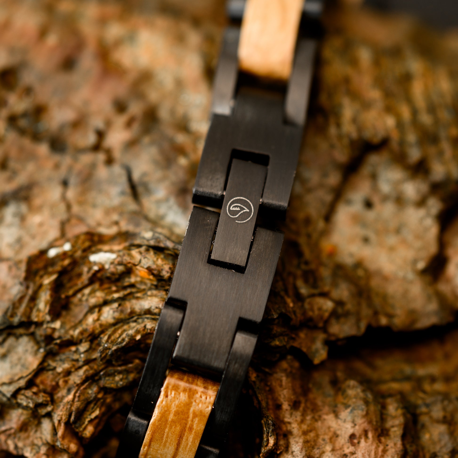 A close up shot of the botanica logo engraved on a black tungsten steel bracelet resting on drift wood
