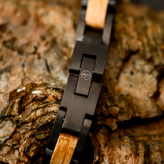 A close up shot of the botanica logo engraved on a black tungsten steel bracelet resting on drift wood
