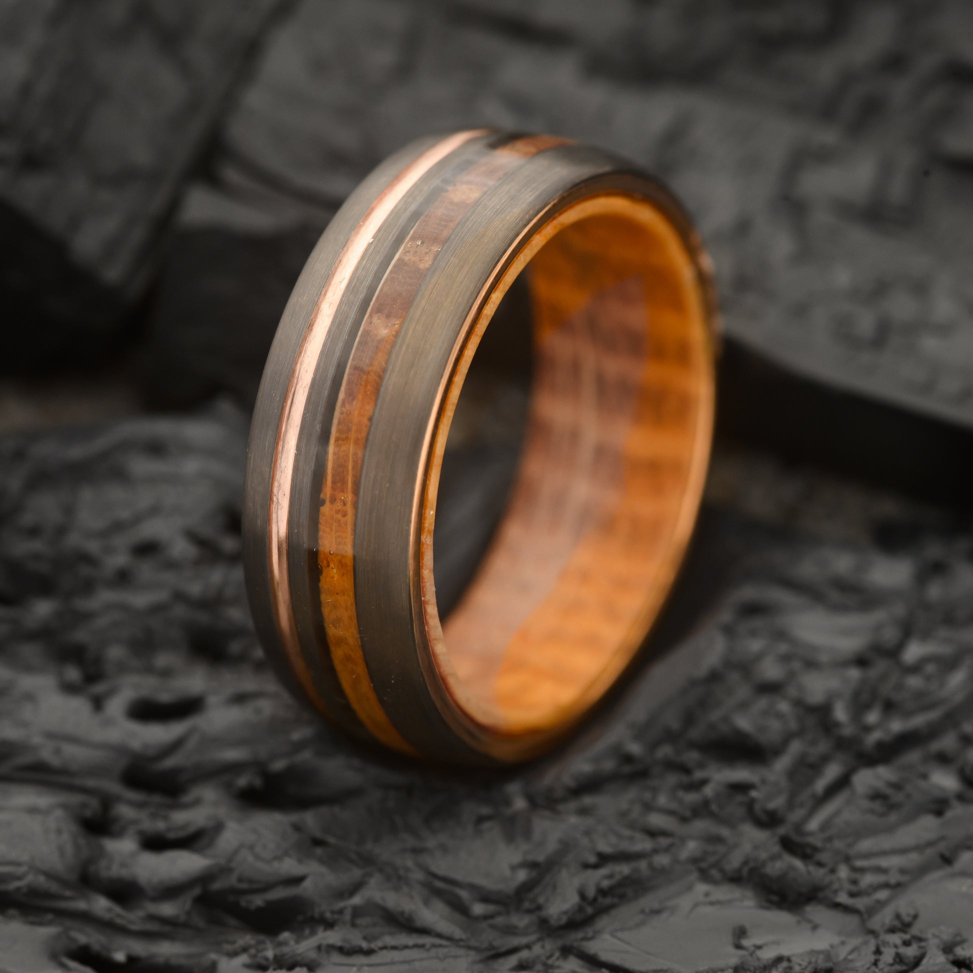 A Mens whiskey wood and rosegold band tungsten steel ring with wood inner ring resting on a charcoal rock