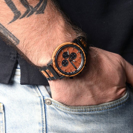 Wooden wristwatch on a person's wrist with tattoos and a denim jacket.