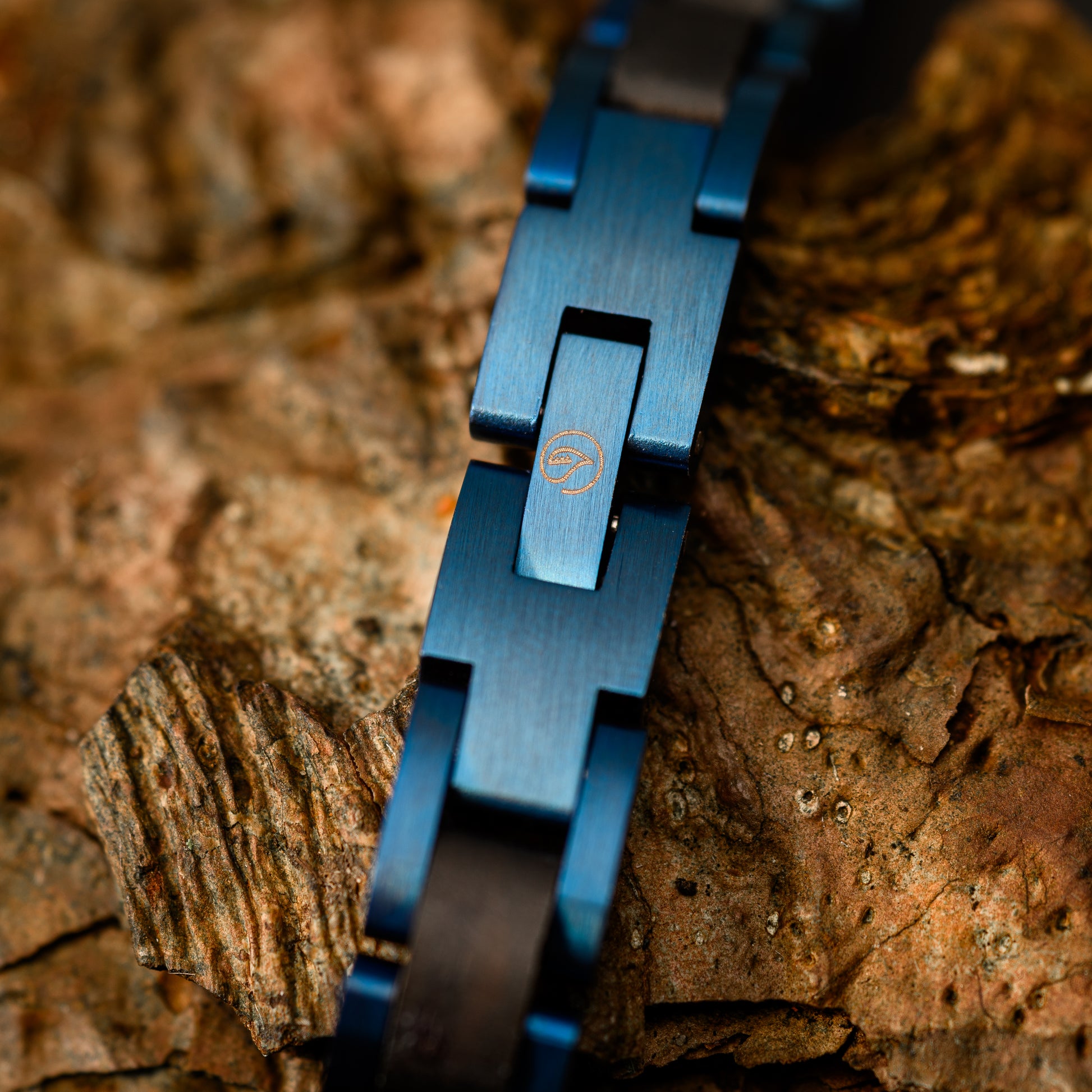 A close up shot of the blue steel bracelet link showing the botanica logo resting on a backdrop of drift wood