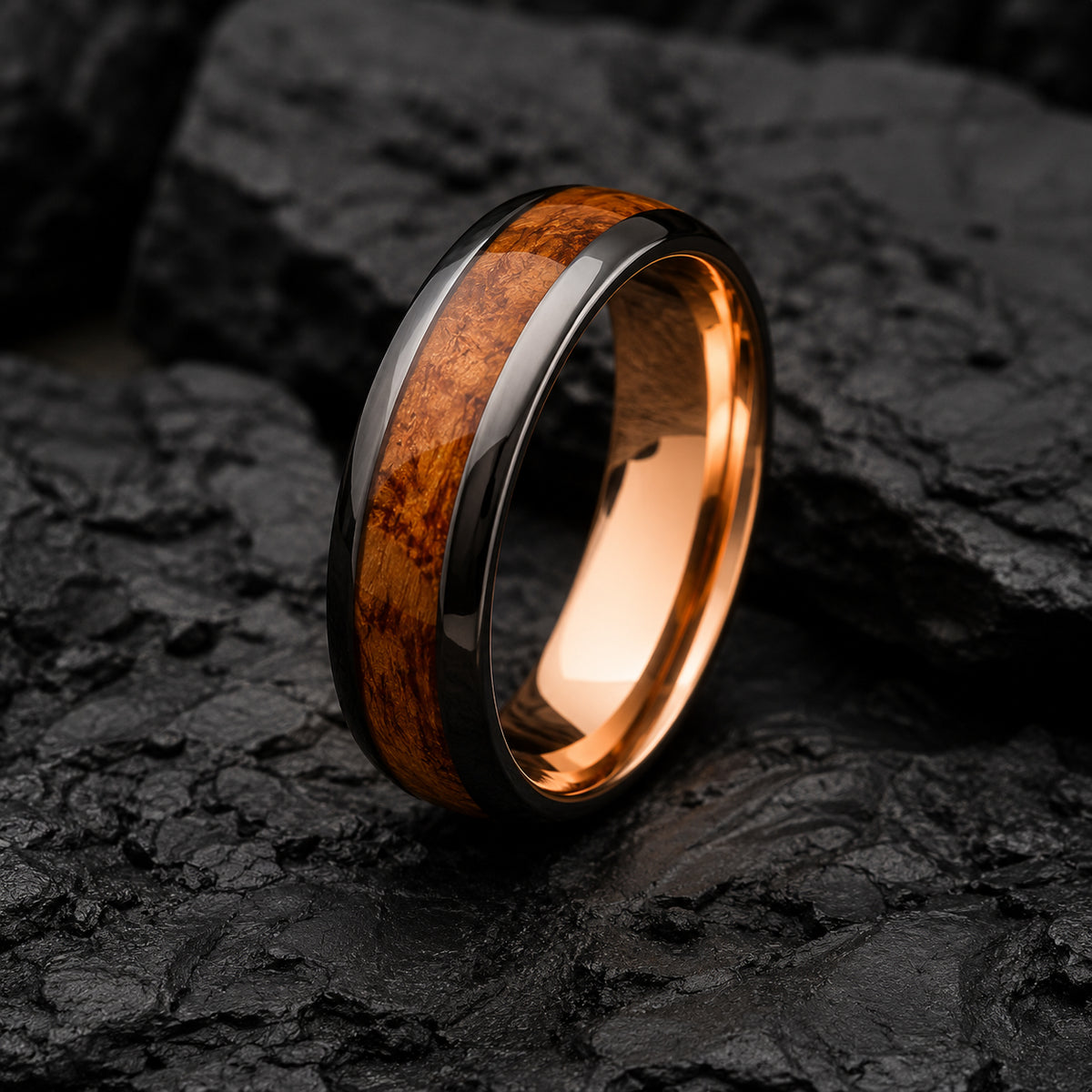 A polished Tungsten ring band with a warm whiskey wood inlay and dark gunmetal gray outer edges, photographed standing upright on dark textured rocks.