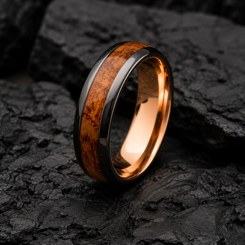 A polished Tungsten ring band with a warm whiskey wood inlay and dark gunmetal gray outer edges, photographed standing upright on dark textured rocks.