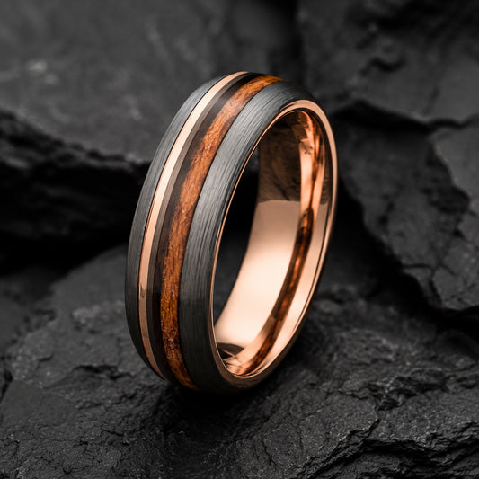 A rose gold and Whiskey wood inlay tungsten ring standing upright on a dark textured slate surface.