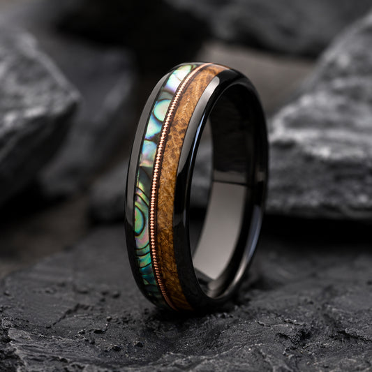 A black Tungsten ring with a double inlay featuring a abalone shell stripe and a Whiskey wood grain stripe, separated by a copper wire border, displayed on dark rocks.