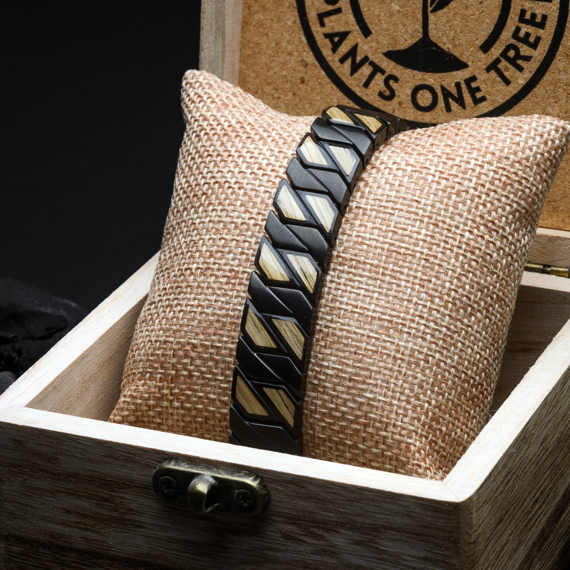 Bracelet with geometric pattern on a textured pillow inside a wooden box, with a brand logo in the background.