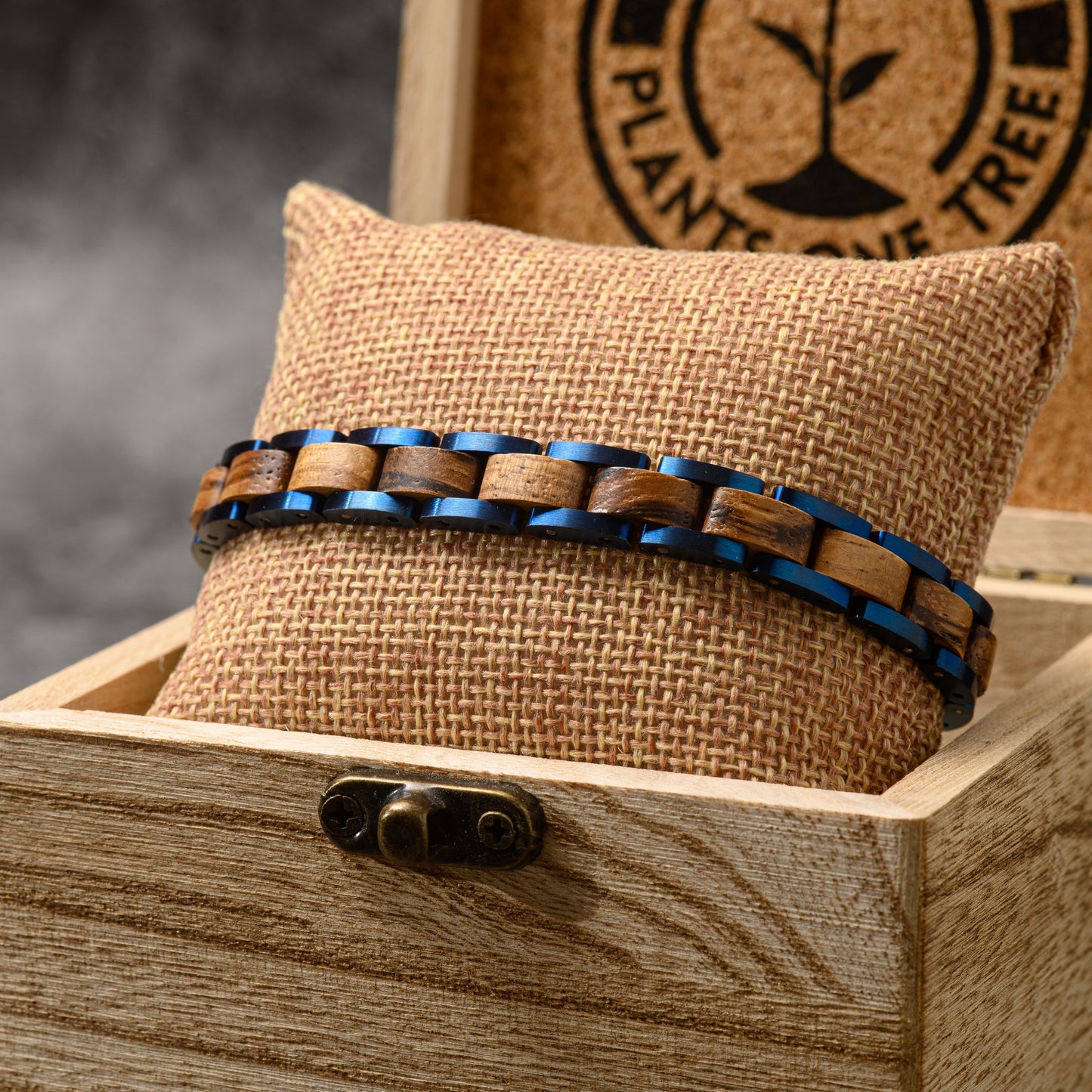 A mens zebrano wood and blue tungsten steel link bracelet resting around a hessian cushion in a wooden presentation box 