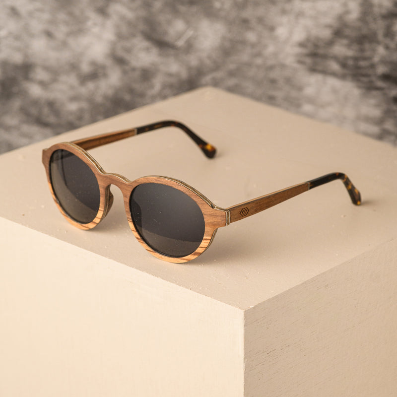 A pair of Lagos wooden sunglasses with a circular frame, featuring layered Zebrano and walnut veneer with aluminium reinforcement. The sunglasses have polarized lenses and are displayed against a neutral background.