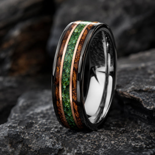 A polished black band tungsten ring with inlaid wood and moss agate, standing on dark textured rocks.