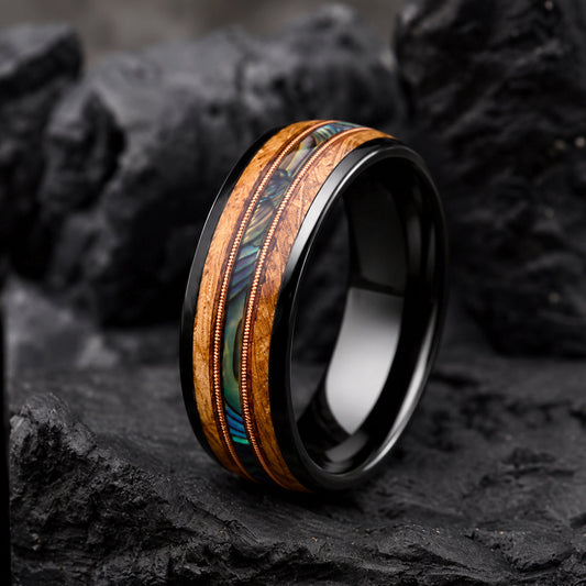 A black Tungsten ring with a central inlay of natural wood and iridescent green-blue abalone shell, displayed on dark textured rocks.